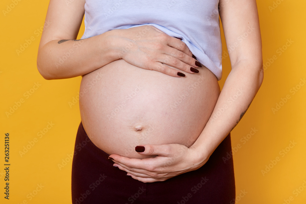Young faceless pregnant woman holds her big belly with hand isolated over yellow background. Pregnant model being photographed in photo studio. Future mother expects baby and posing with bare abdomen. Young faceless pregnant woman holds her big belly with hand isolated over yellow background. Pregnant model being photographed in photo studio. Future mother expects baby and posing with bare abdomen.