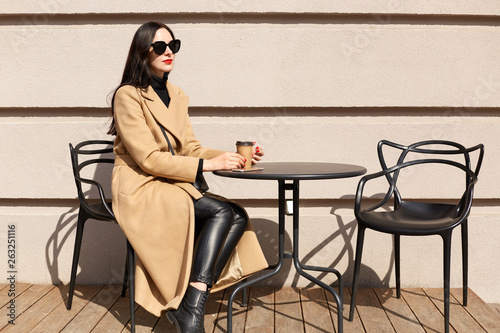 Pleased beautiful young model poses isolated sitting over light wall on terrace, having coffee break, enjoys sunny day, looking aside. Slender pretty female wearing stylish beige coat and black pants.