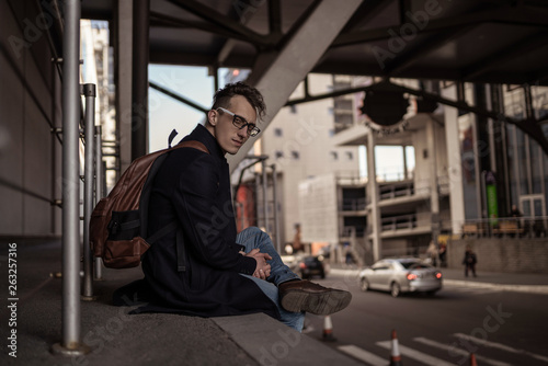 Wallpaper Mural Young man in black coat and glasses with backpack sitting on city street near shopping center. Street style photo shoot. Torontodigital.ca