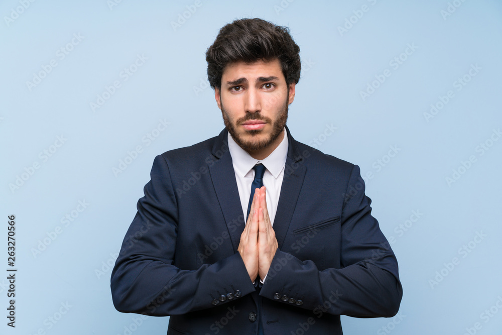 Businessman over isolated blue wall keeps palm together. Person asks for something Businessman over isolated blue wall keeps palm together. Person asks for something