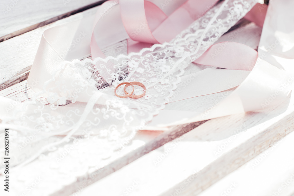 Gold wedding rings with pink lace and ribbons lie on a white wooden surface. Gold wedding rings with pink lace and ribbons lie on a white wooden surface.