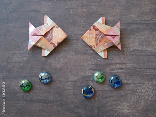 Two origami fish made of ten euro notes facing each other on dark wooden table area