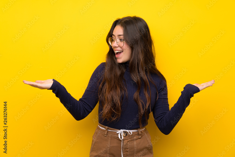 Teenager girl over isolated yellow wall holding copyspace with two hands Teenager girl over isolated yellow wall holding copyspace with two hands