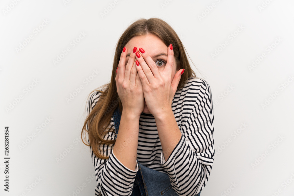 Young woman in dungarees over white wall covering eyes and looking through fingers Young woman in dungarees over white wall covering eyes and looking through fingers