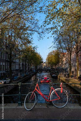 Red bicycle parking on the bridge over the calm water of a canal with a boat giving a round trip to tourists, Amsterdam, Netherlands