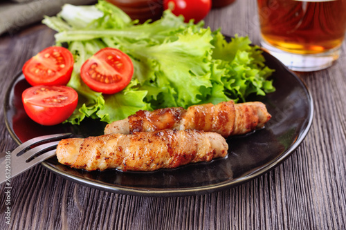 Appetizing sausages wrapped in bacon and grilled next to tomatoes, lettuce and and glass of beer are on a wooden table