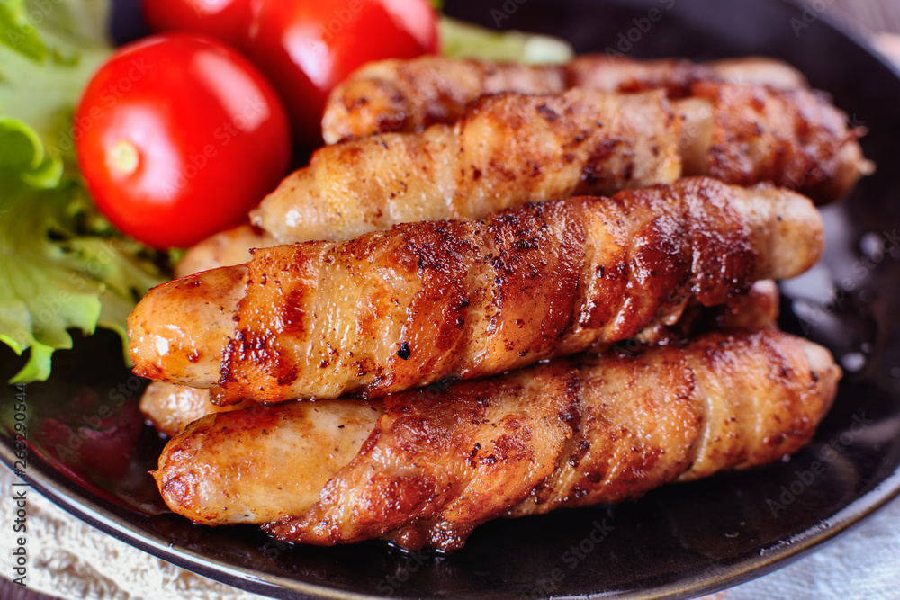 Appetizing sausages wrapped in bacon and grilled next to tomatoes and lettuce are on a wooden table, macro