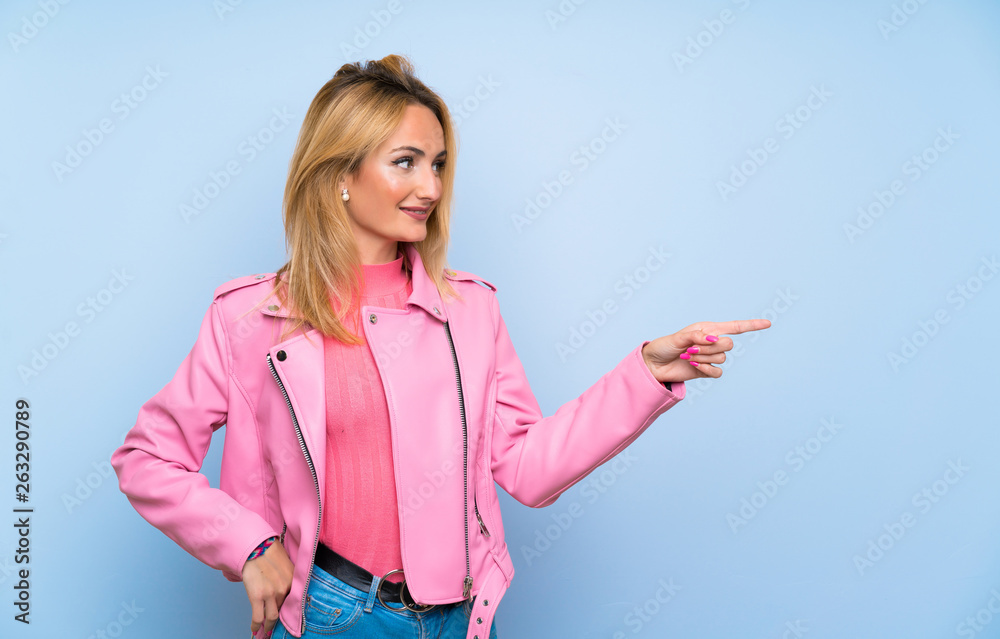 Young blonde woman with pink jacket over isolated blue background pointing finger to the side Young blonde woman with pink jacket over isolated blue background pointing finger to the side