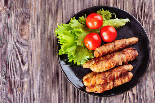 Appetizing sausages wrapped in bacon and grilled next to tomatoes and lettuce are on a wooden table, top view