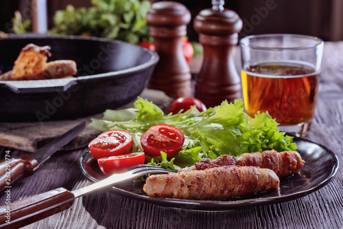 Appetizing sausages wrapped in bacon and grilled next to tomatoes and lettuce are on a wooden table