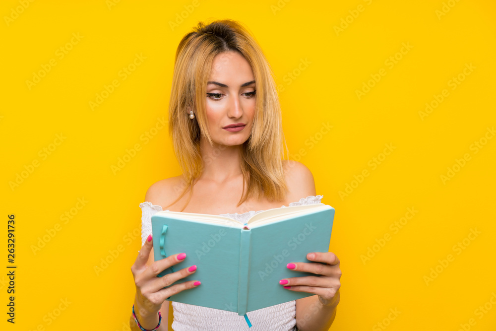 Young blonde woman over isolated yellow wall holding and reading a book Young blonde woman over isolated yellow wall holding and reading a book