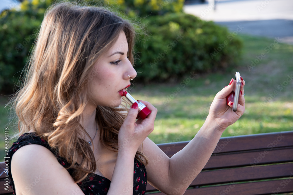 Portrait of a blonde girl who is putting makeup on red lips and using her phone as a mirror. Natural background with tree. Copy space.