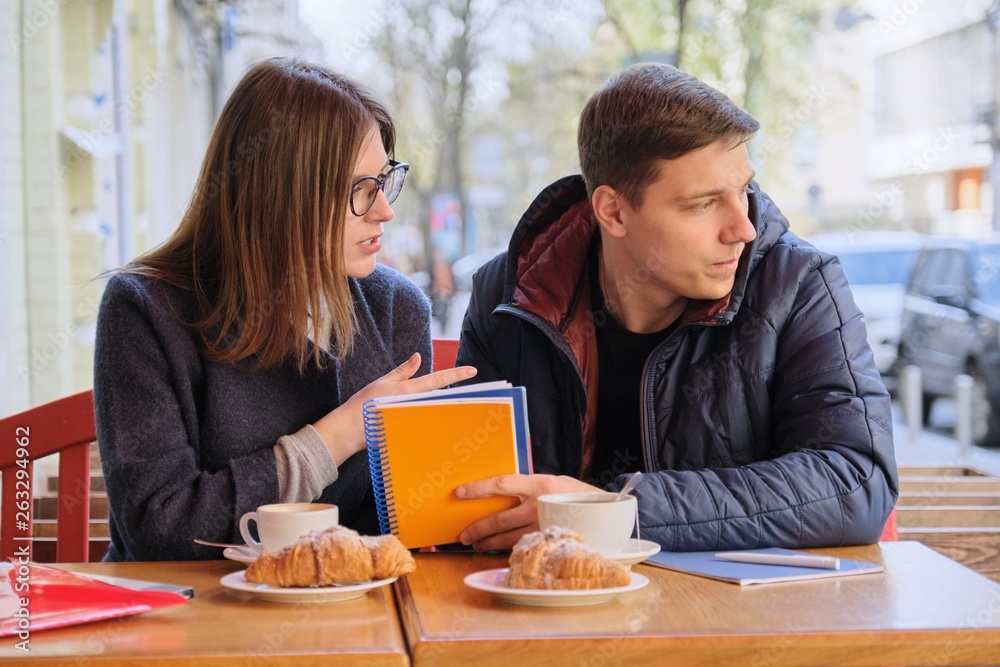 Fototapeta premium Young couple of students study in outdoor cafe, drink coffee tea, eat croissants, background is spring city street