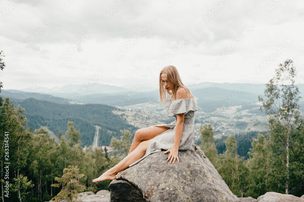 Naklejka premium Young beautiful long hair blonde sitting at stone on mountain with picturesque view at hiils and cloudy sky. Cute woman in summer dress relaxing and posing outdoor portrait. Happy female at nature.