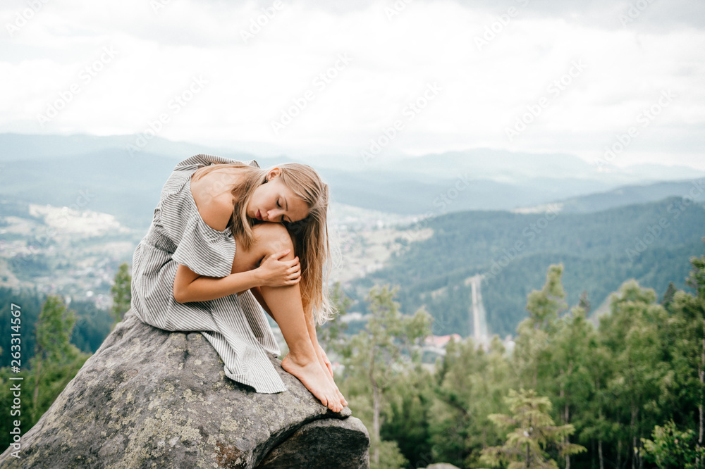 Lonely young girl sits at stone on top of mountain with far view at hills and cloudy sky and hugs her legs with head lying on knees. Beautiful blonde woman outdoor summer  portrait. Female at nature.