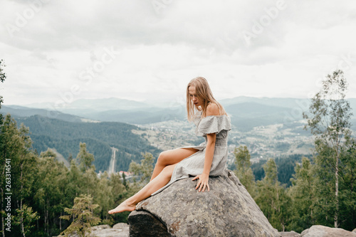 Young beautiful long hair blonde sitting at stone on mountain with picturesque view at hiils and cloudy sky. Cute woman in summer dress relaxing and posing outdoor portrait. Happy female at nature.
