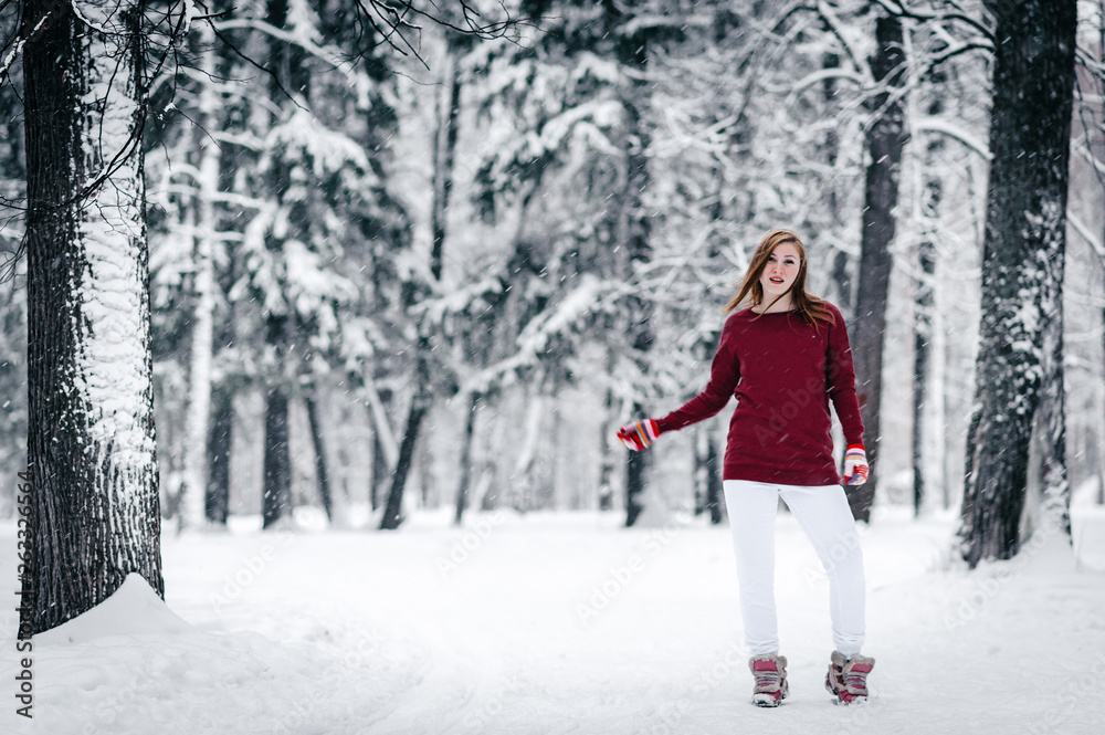 the girl dressed in a maroon sweater and white pants stands against the tree trunk against a backdrop of snow-covered winter forest