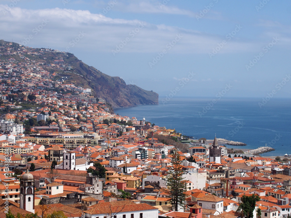 a sunlit view of the city of funchal from above with rooftops and buildings in front of a bright blue sea