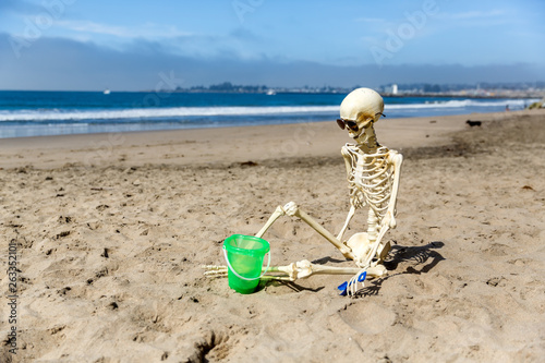 Skeleton sits on the beach playing in the sand with a bucket and shovel photo