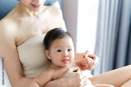 Asian cute baby sitting on the lap of her mother on the bed. Baby is looking on the camera while mother holding baby's body and arm and looking with smile. Touch of love from mother concept.