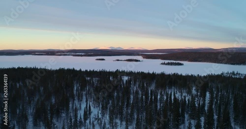 Aerial, reverse, drone shot, over spruce forest, buildings and Ounastunturit fjells, in Pallas-yllastunturi national park, at the arctic circle, on a sunny, winter evening, in Muonio, Lapland, Finland photo