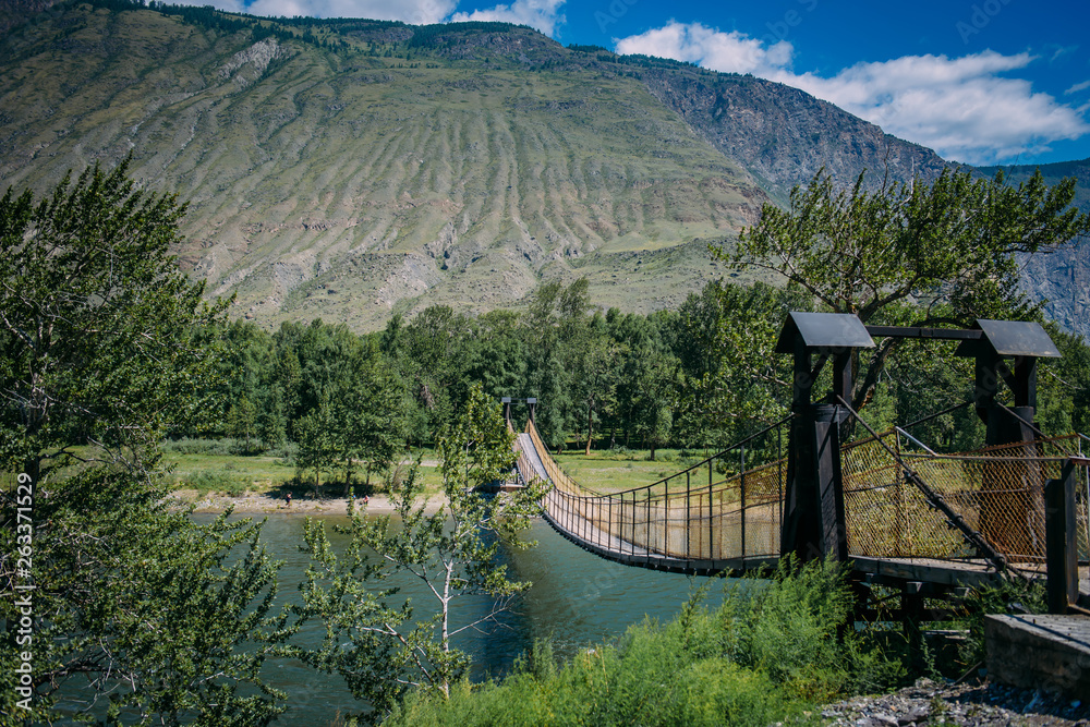Suspension bridge over the mountain river. The hanging bridge surrounded by lush green trees on the background of mountains and white clouds on the blue sky. Travelling to Russia.
