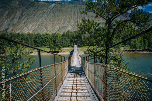 Fototapeta Naklejka Na Ścianę i Meble -  Suspension bridge over the mountain river. The hanging bridge surrounded by lush green trees on the background of mountains and white clouds on the blue sky. Travelling to Russia.