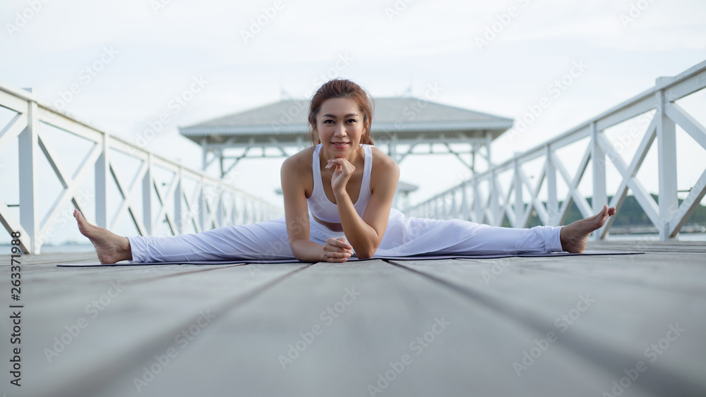 Happy young woman in white clothes practicing yoga. doing splits. stretching in Samakonasana exercise..enjoying sea view, concept for exercising, health care