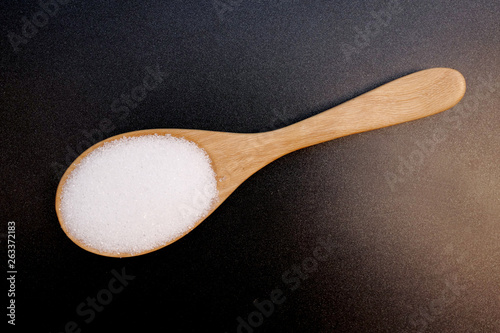 White sugar on wooden spoon on black background, top view.