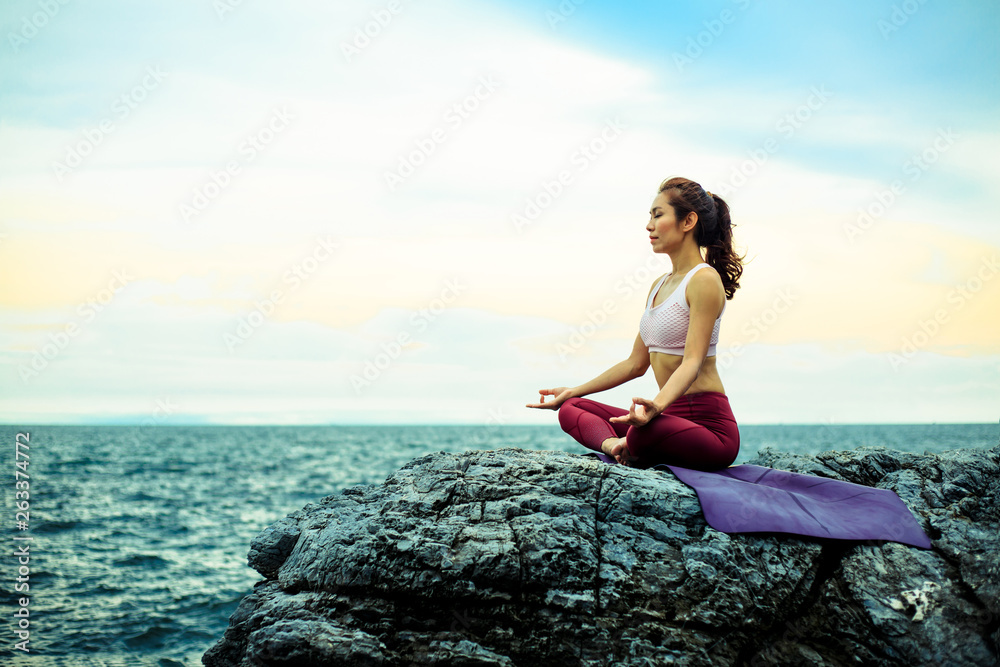 Young woman doing exercise yoga. sitting on the rock, doing Padmasana exercise, Lotus pose. Amazing yoga landscape in beautiful sky and enjoying sea view with evening.