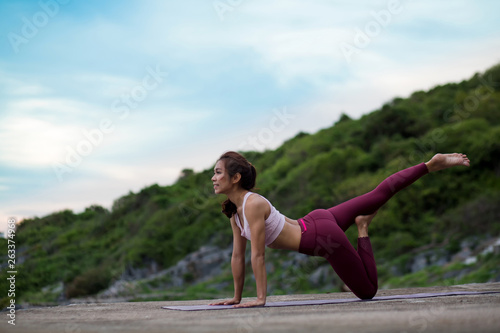 Side view of Women practicing flexible yoga pose. smiling woman stretching leg on mat on the rock. Amazing yoga landscape and and enjoying outdoors mountains view , concept for exercising, health care