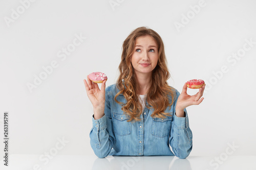 Confused young pretty woman with long blond wavy hair, sitting at the table and holds donuts in her hands. Looks up and wonders whether to eat sweets for the night. isolated over white background.