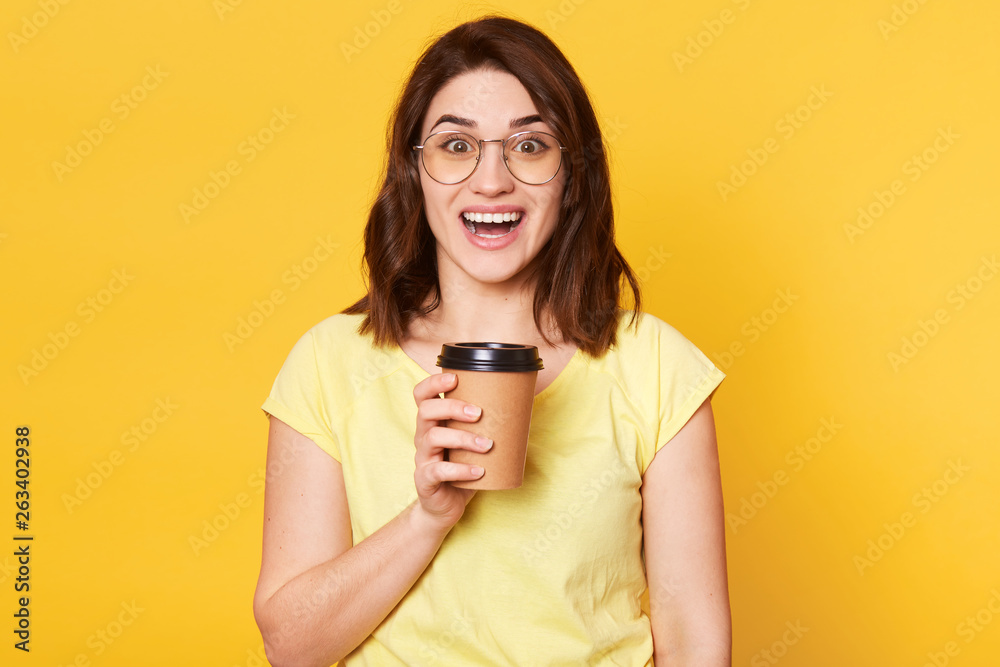 Close up portrait of young lady isolated on yellow studio background, posing with happy facial expression while drinking tekeaway coffee, has positive feelings and toothy smile, stands with open mouth Close up portrait of young lady isolated on yellow studio background, posing with happy facial expression while drinking tekeaway coffee, has positive feelings and toothy smile, stands with open mouth