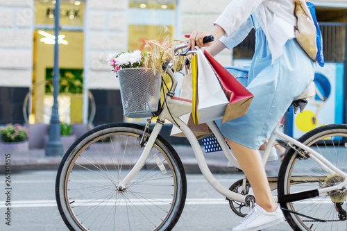 Young woman is cycling around city on white bicycle. Girl in blue dress is going shopping with colorful packages on handlebars of bike. Female retro bicycle with basket with bouquet of flowers.