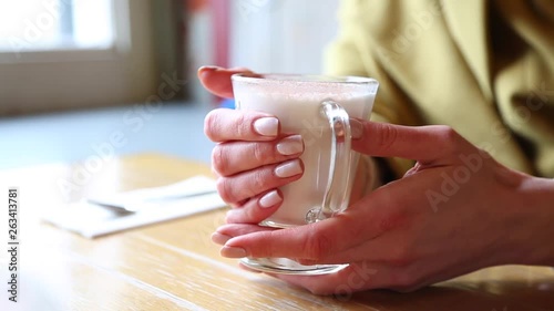 A glass beaker with hot foamed milk. Woman warms hands on a glass mug of hot milk in a cafeshop. Beautiful latte with a big foam. A woman touches a glass with a hot latte