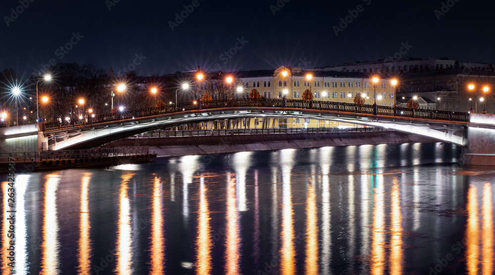 Night bridge with lanterns in the form of stars across the river, which reflects the lights of the big city
