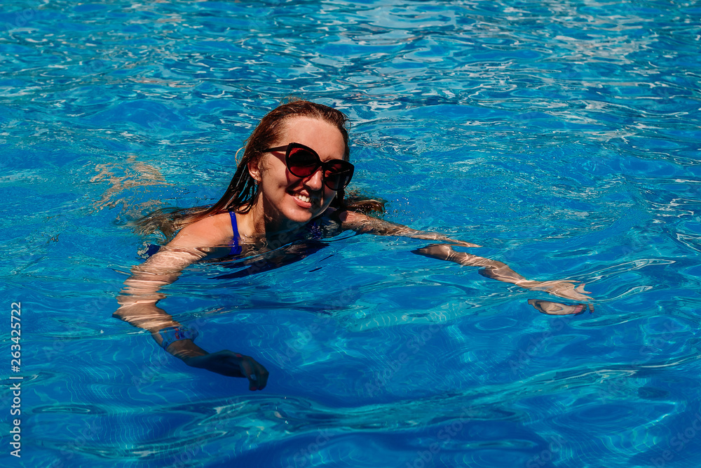 a smiling young woman in a swimsuit and sunglasses is swimming in an outdoor pool with blue water .