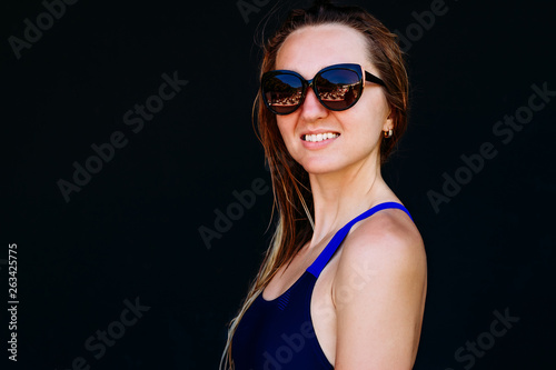 smiling tanned woman in a sports swimsuit and sunglasses stands on a black background. Female portrait