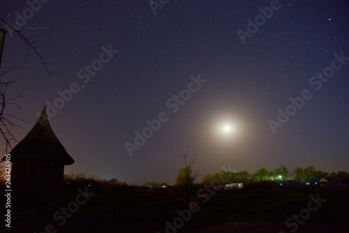 Evening shining starry sky  blue space background with stars. Night sky full of stars and sunrise on the background horizon. Purple dark sky with many stars above field of trees  house and moon