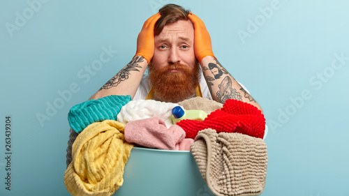 Fotografie Troubled ginger young man keeps hands on head, looks desperately, wears protective gloves, has tattooed arms, sorrowful facial expression, stands against blue background