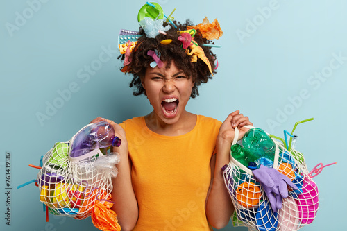 Irritated black young woman collects plastic waste  keeps mouth opened  holds net bags with litter  expresses negative emotions  demands save nature  recycles garbage. Problem of environment