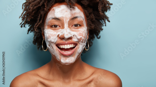 Happy Afro American female laughs positively, has white even teeth, washes face with soap, cleanses skin, stands half naked against blue background. Women, beauty, facial treatments concept.