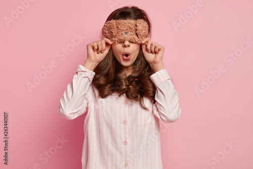 Surprised beautiful lady covers eyes with sleepmask, wears casual pyjamas, opens mouth, has curly hair, isolated over pink background, prepares for sleep, poses in bedroom. Sleeping concept. © Wayhome Studio
