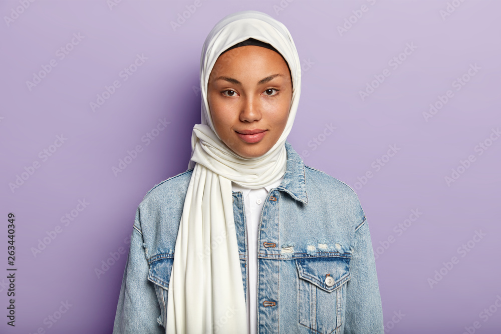 Beautiful woman with healthy dark skin, wears white hijab, dressed in denim jacket, looks directly at camera, isolated over purple background. Religious female has natural beauty. Ethnicity concept Beautiful woman with healthy dark skin, wears white hijab, dressed in denim jacket, looks directly at camera, isolated over purple background. Religious female has natural beauty. Ethnicity concept