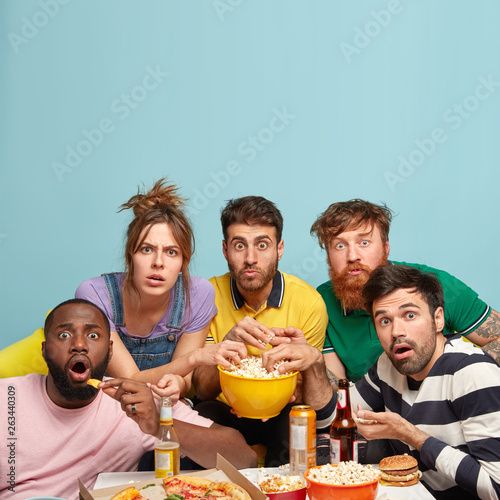 Vertical shot of shocked mixed race companions or fellows, watch terror movie with popcorn, have scared expressions, eat crisps and pizza, isolated over blue background with freee space above photo