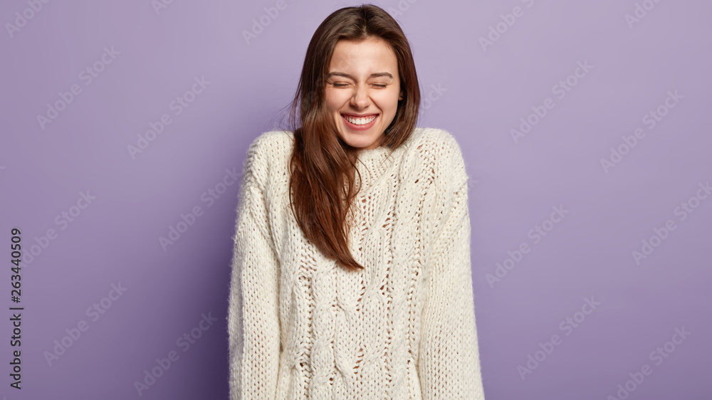 Vivacious overjoyed girl with beaming smile, laughs from pleasure, has white teeth, dressed in long sleeve sweater, closes eyes, has dark hair, models over purple studio wall. Positive emotions Vivacious overjoyed girl with beaming smile, laughs from pleasure, has white teeth, dressed in long sleeve sweater, closes eyes, has dark hair, models over purple studio wall. Positive emotions