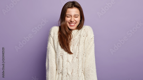Vivacious overjoyed girl with beaming smile, laughs from pleasure, has white teeth, dressed in long sleeve sweater, closes eyes, has dark hair, models over purple studio wall. Positive emotions