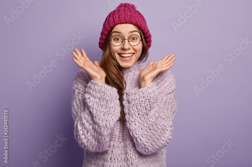 Fotografie Photo of happy emotive European woman gestures with both hands, clasps palms, wears round spectacles, dressed in winter hat, knitted jumper, isolated over purple background