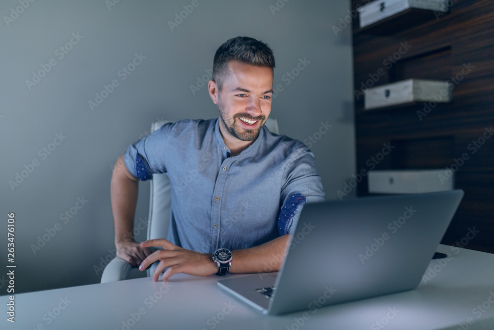 Smiling hardworking Caucasian businessman looking at laptop while sitting in office late at night. Smiling hardworking Caucasian businessman looking at laptop while sitting in office late at night.