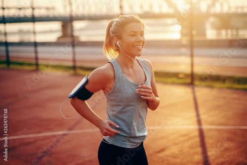 Charming female blonde runner in sportswear running on court with earphones in ears and smart phone in phone case around arm. Morning fitness concept. Running slow is not character flaw, quitting is.